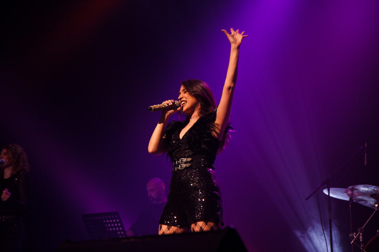 Olive Shows Madonna tribute - the soloist in a black feathered sequin dress reaching one arm high while singing into a jewelled microphone under purple light